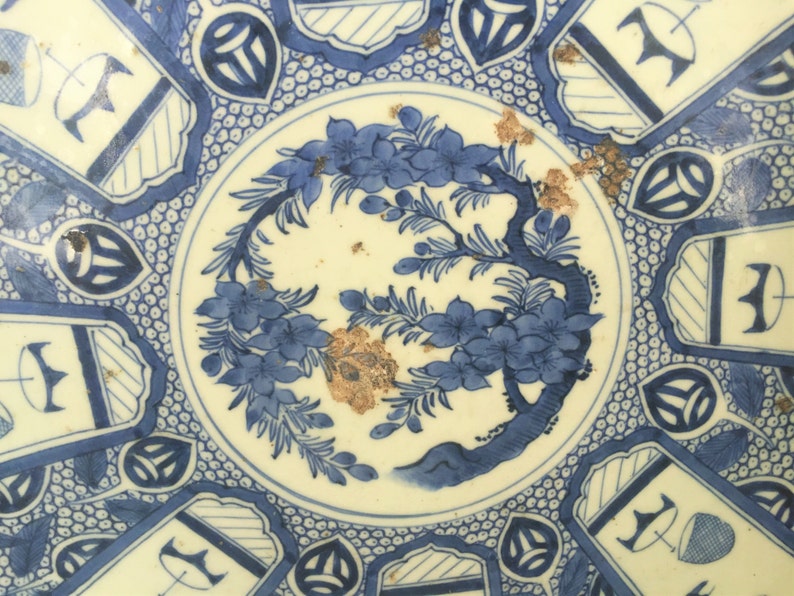 Op de afbeelding: Een blauw en wit keramisch bord met een bloemenontwerp en geometrische patronen. Het bord heeft een centraal ontwerp van een boom met bloemen en bladeren, omringd door een rand van zich herhalende geometrische vormen.