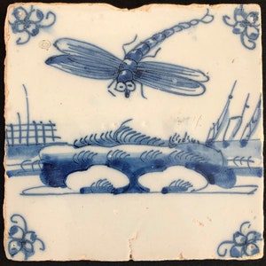 Puede incluir: Azulejo cer&aacute;mico cuadrado blanquecino con un dise&ntilde;o de lib&eacute;lula azul. La lib&eacute;lula est&aacute; sobre un paisaje estilizado con un puente y vegetaci&oacute;n. Acentos florales en las esquinas completan el dise&ntilde;o. El azulejo tiene un aspecto ligeramente envejecido.