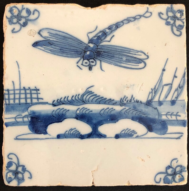 Puede incluir: Azulejo cer&aacute;mico cuadrado blanquecino con un dise&ntilde;o de lib&eacute;lula azul. La lib&eacute;lula est&aacute; sobre un paisaje estilizado con un puente y vegetaci&oacute;n. Acentos florales en las esquinas completan el dise&ntilde;o. El azulejo tiene un aspecto ligeramente envejecido.