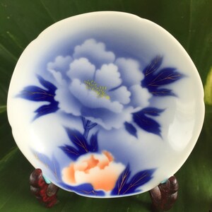 May include: A small, white ceramic dish featuring a blue peony design with gold detailing. The dish is supported by a small, dark wooden stand. The background is a large green leaf. The dish's diameter is approximately 10 cm.