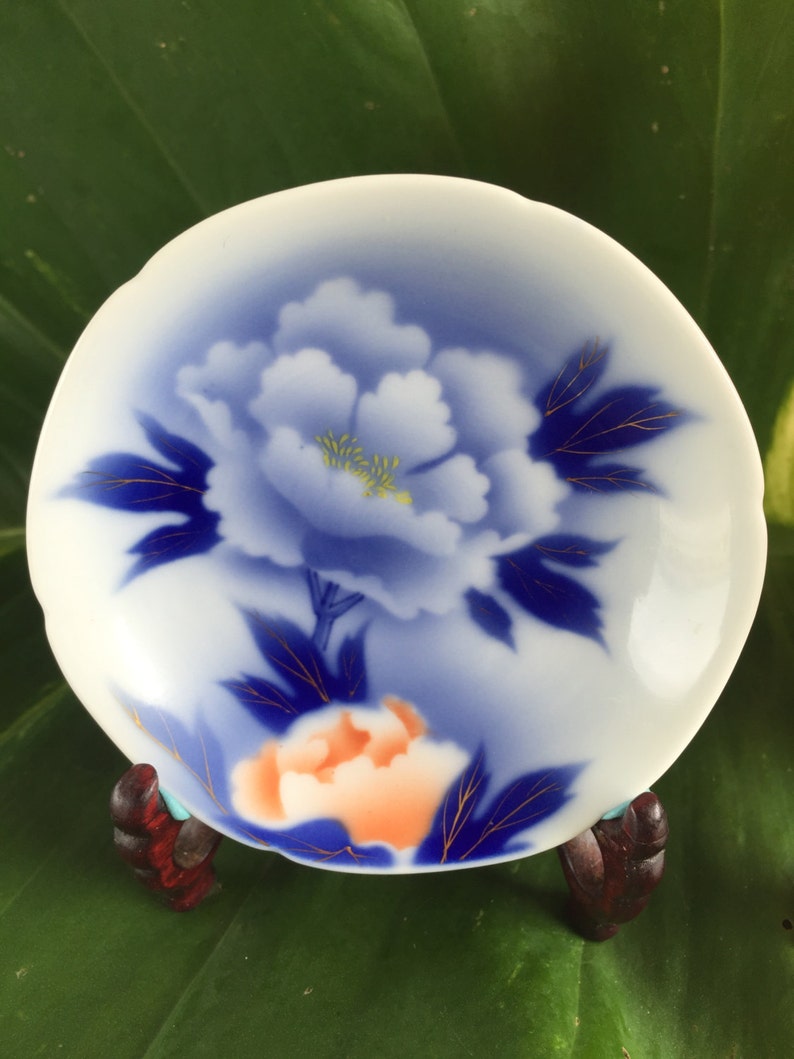 May include: A small, white ceramic dish featuring a blue peony design with gold detailing. The dish is supported by a small, dark wooden stand. The background is a large green leaf. The dish's diameter is approximately 10 cm.