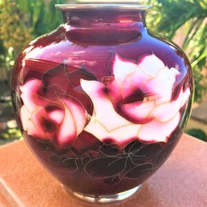 Puede incluir: Un jarr&oacute;n de cer&aacute;mica rojo oscuro con un borde plateado y un dise&ntilde;o floral de rosas blancas y rosas con detalles dorados.