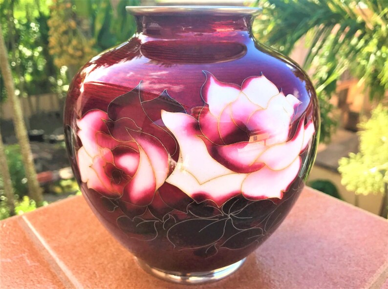 Puede incluir: Un jarr&oacute;n de cer&aacute;mica rojo oscuro con un borde plateado y un dise&ntilde;o floral de rosas blancas y rosas con detalles dorados.