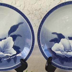 Op de afbeelding: Twee bijpassende keramische borden met een wit en blauw bloemmotief. De borden hebben een witte bloem in het midden, omringd door blauwe bladeren en een blauwe rand. De borden worden op zwarte standaards gepresenteerd.