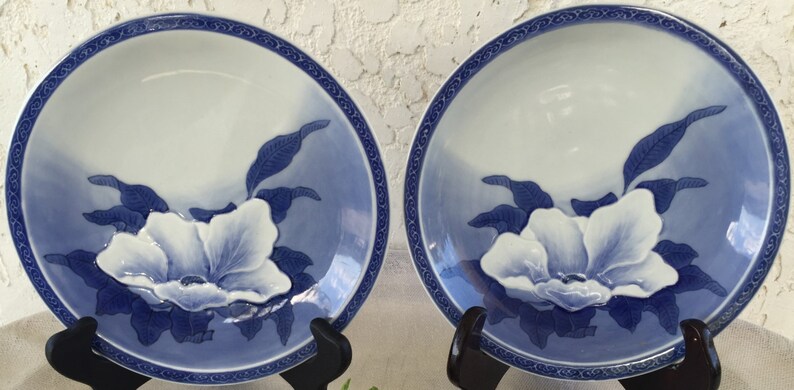 Op de afbeelding: Twee bijpassende keramische borden met een wit en blauw bloemmotief. De borden hebben een witte bloem in het midden, omringd door blauwe bladeren en een blauwe rand. De borden worden op zwarte standaards gepresenteerd.