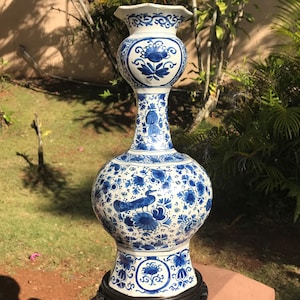 May include: A tall, ornate blue and white ceramic vase with a bulbous body and a flared top. The vase features intricate floral and bird designs. It stands on a dark wooden base and is approximately 91 cm tall.