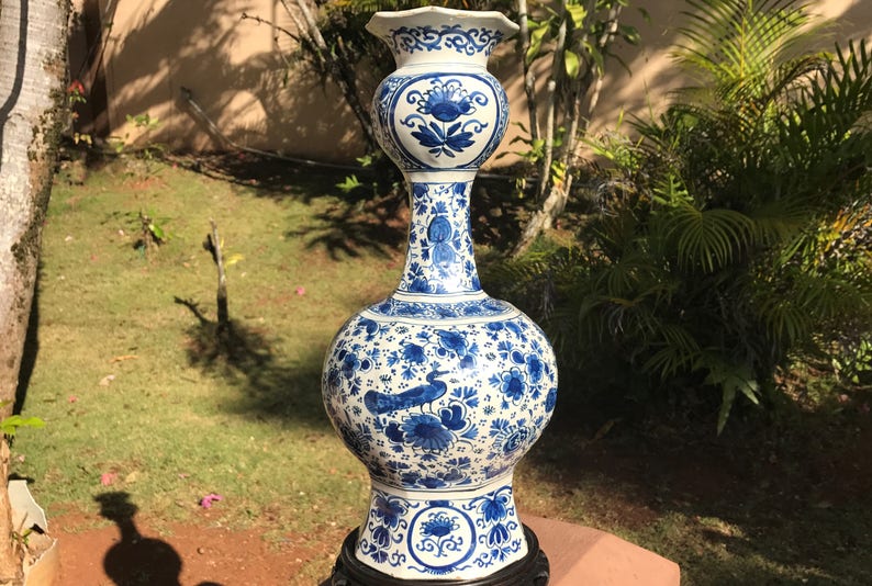 May include: A tall, ornate blue and white ceramic vase with a bulbous body and a flared top. The vase features intricate floral and bird designs. It stands on a dark wooden base and is approximately 91 cm tall.