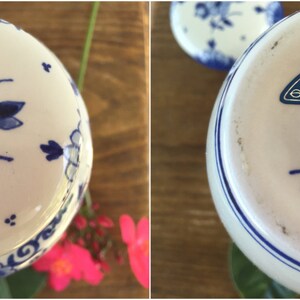 May include: A white ceramic vase with blue floral designs and a blue rim. The bottom of the vase has a blue stamp that reads "KONINKLIJKE PORCELEYNE FLES" and other text. The vase is on a wooden surface.