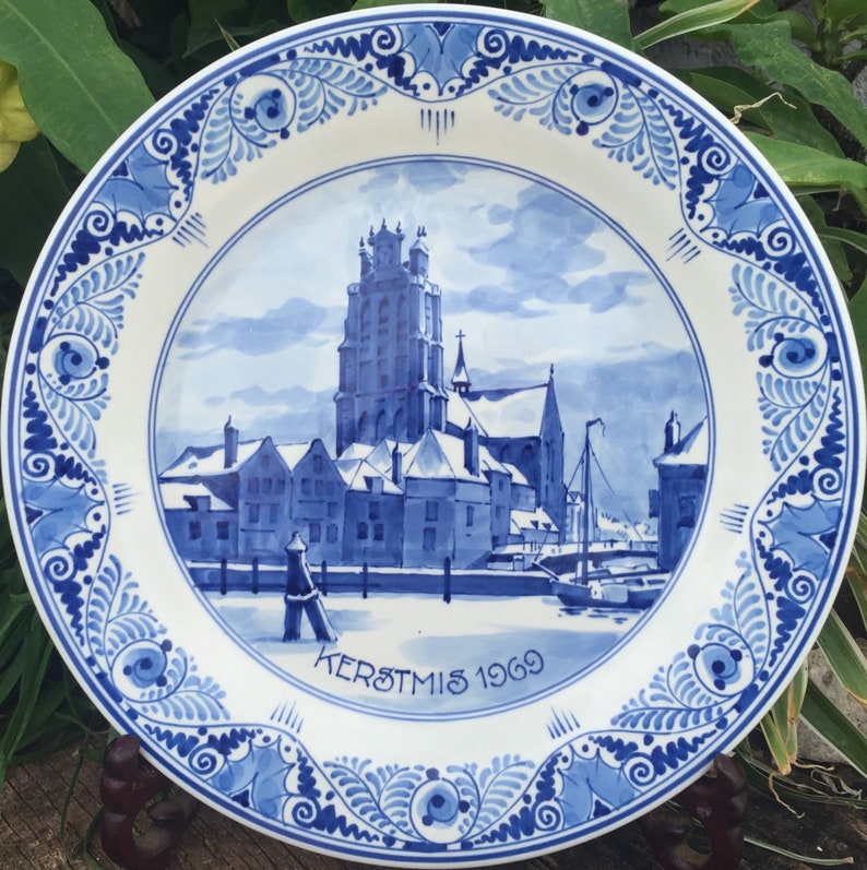 May include: A white ceramic plate with a blue and white painted design. The plate features a detailed illustration of a church and buildings, with the text "KERSTMIS 1969". The border has a decorative floral pattern.