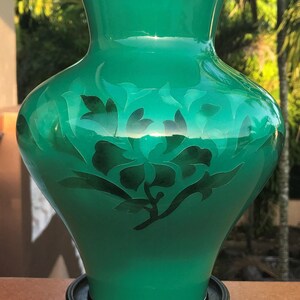 May include: A vivid green vase with a floral pattern, presented on a dark wooden stand. The vase has a shiny finish and a silver rim. The floral design is a darker green, offering a striking contrast.