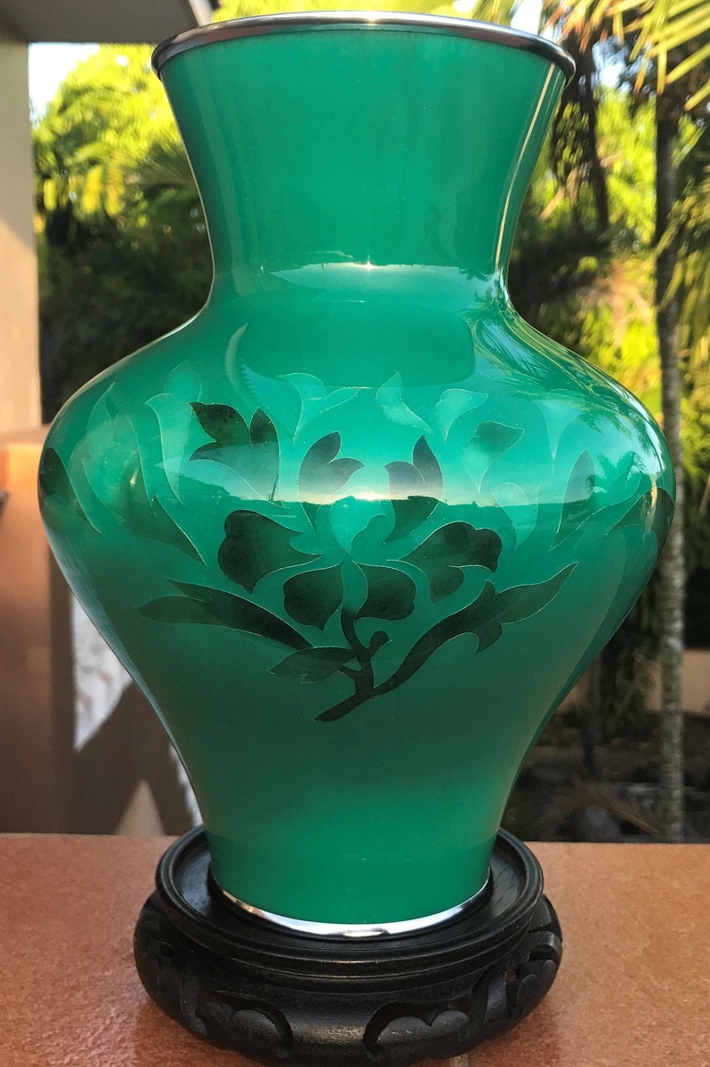 May include: A vivid green vase with a floral pattern, presented on a dark wooden stand. The vase has a shiny finish and a silver rim. The floral design is a darker green, offering a striking contrast.