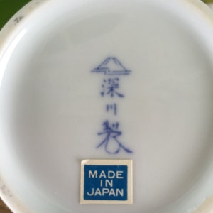 Może przedstawiać: Zbliżenie na sp&oacute;d białego ceramicznego przedmiotu z niebieskimi japońskimi znakami i niebiesko-białą naklejką "MADE IN JAPAN". Obiekt ma lekko zakrzywioną krawędź.