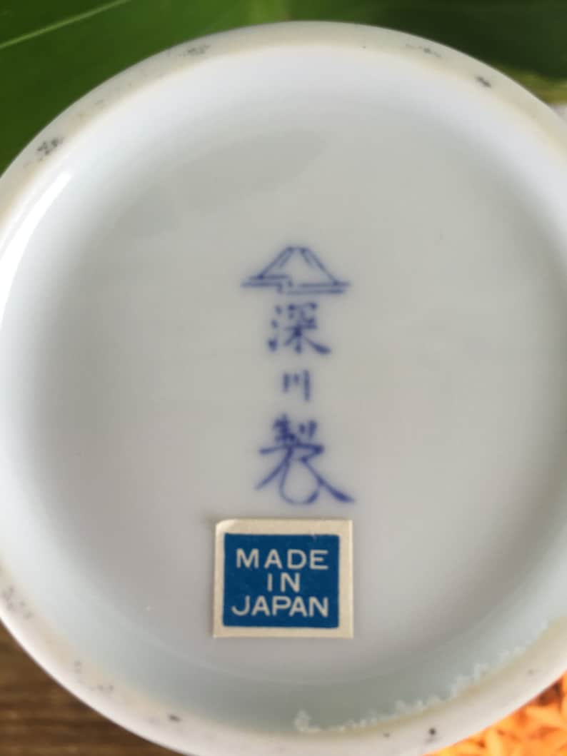 Może przedstawiać: Zbliżenie na sp&oacute;d białego ceramicznego przedmiotu z niebieskimi japońskimi znakami i niebiesko-białą naklejką "MADE IN JAPAN". Obiekt ma lekko zakrzywioną krawędź.