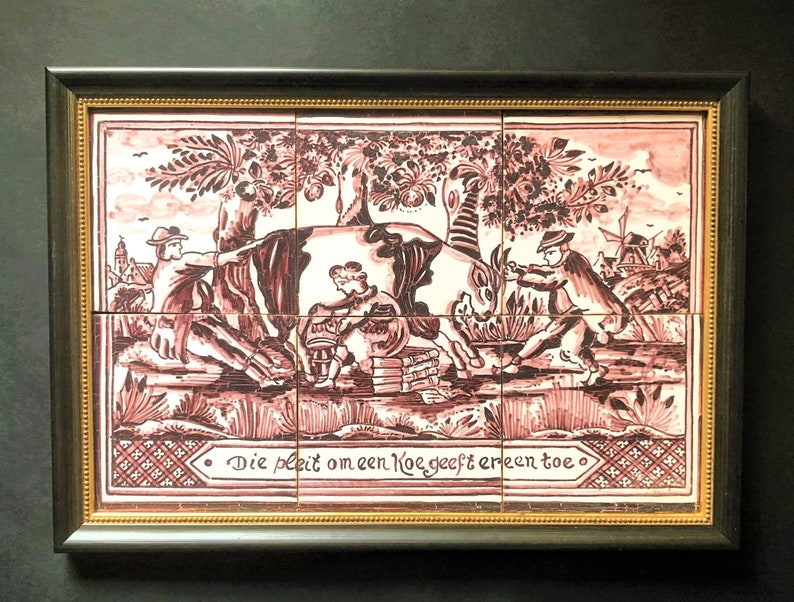 May include: A framed tile depicting a scene of people interacting with a cow. The tile is decorated with a pink and white floral pattern and the text "Die pleit om een koe geeft er een toe".