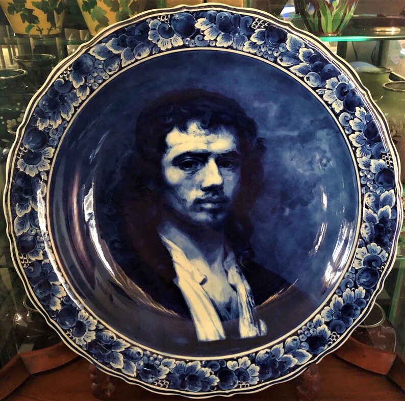 May include: A large, round, blue and white decorative plate featuring a portrait of a man. The plate has a scalloped edge and a floral border. The portrait is painted in shades of blue and white.