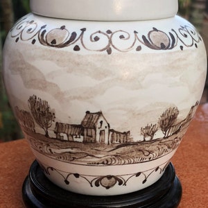 Puede incluir: Jarra de jengibre de cer&aacute;mica blanca con tapa, con una escena de paisaje pintada a mano en tonos marrones. La jarra se asienta sobre un soporte de madera negro. La parte superior e inferior est&aacute;n bordeadas con volutas decorativas.
