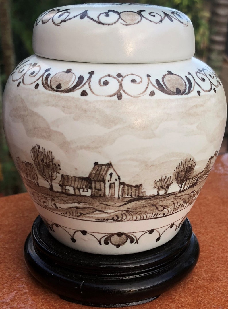 Puede incluir: Jarra de jengibre de cer&aacute;mica blanca con tapa, con una escena de paisaje pintada a mano en tonos marrones. La jarra se asienta sobre un soporte de madera negro. La parte superior e inferior est&aacute;n bordeadas con volutas decorativas.