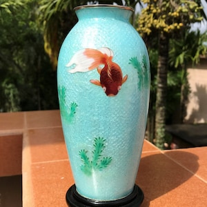 Pu&ograve; includere: Un vaso in ceramica blu turchese con un pesce dorato e piante verdi dipinte su di esso. Il vaso ha una superficie testurizzata e si trova su una base in legno nero.