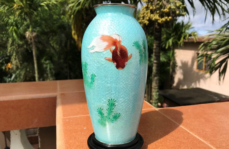Pu&ograve; includere: Un vaso in ceramica blu turchese con un pesce dorato e piante verdi dipinte su di esso. Il vaso ha una superficie testurizzata e si trova su una base in legno nero.