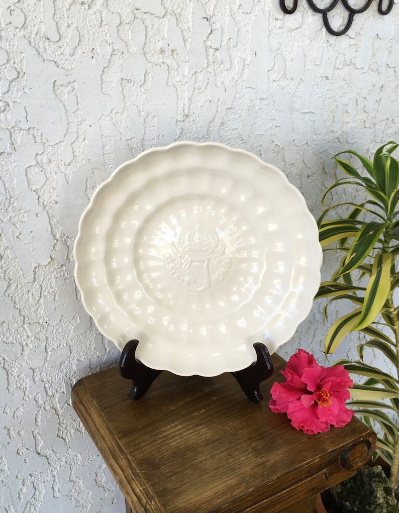 Puede incluir: Un plato de cer&aacute;mica blanco con un borde festoneado y un dise&ntilde;o en relieve en el centro. El plato est&aacute; sobre un soporte de madera.