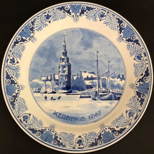 May include: A blue and white ceramic plate depicting a Dutch winter scene. The plate features a church, houses, and a canal with people ice skating. The text "KERSTMIS 1967" is written on the plate.