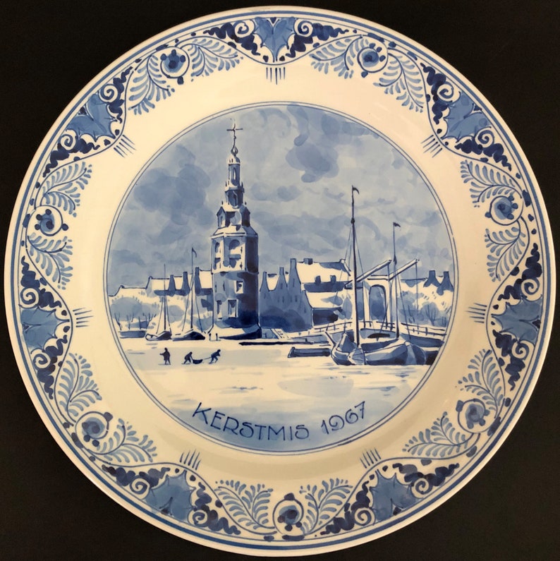 May include: A blue and white ceramic plate depicting a Dutch winter scene. The plate features a church, houses, and a canal with people ice skating. The text "KERSTMIS 1967" is written on the plate.