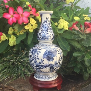 Könnte beinhalten: Eine weiß-blaue Keramikvase mit detailliertem Vogel- und Blumendesign. Die Vase steht auf einem dunkelbraunen Holzsockel. Der Hintergrund zeigt grünes Laub und rosa und gelbe Blüten.