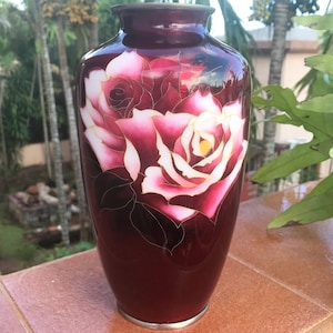 Puede incluir: Un jarr&oacute;n de cer&aacute;mica rojo oscuro con un dise&ntilde;o de rosa blanca y rosa. El jarr&oacute;n est&aacute; decorado con detalles dorados.