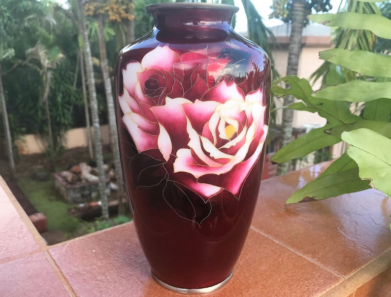 Puede incluir: Un jarr&oacute;n de cer&aacute;mica rojo oscuro con un dise&ntilde;o de rosa blanca y rosa. El jarr&oacute;n est&aacute; decorado con detalles dorados.