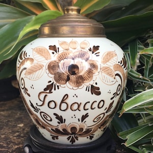 May include: A white ceramic tobacco jar with a brass lid, adorned with brown floral patterns and the word "Tobacco" in elegant script. The jar is set against a backdrop of lush green foliage.