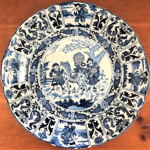 May include: A large, round, blue and white decorative plate with a scalloped edge. The plate features a central scene of figures with a horse, surrounded by floral and foliate patterns. The overall design is reminiscent of antique porcelain.
