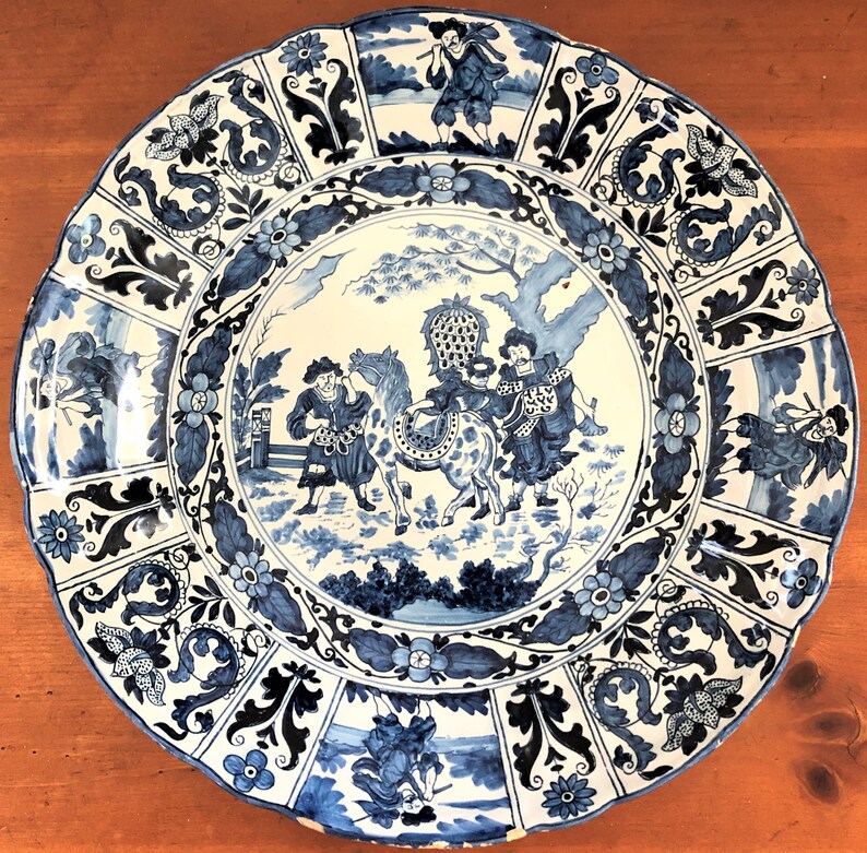 May include: A large, round, blue and white decorative plate with a scalloped edge. The plate features a central scene of figures with a horse, surrounded by floral and foliate patterns. The overall design is reminiscent of antique porcelain.