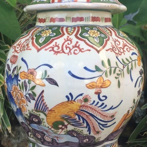May include: A decorative ceramic jar with a lid, featuring a vibrant floral and bird design. The jar is predominantly white, adorned with colorful flowers, birds, and intricate patterns in shades of blue, yellow, red, and green. It sits on a dark wooden base.