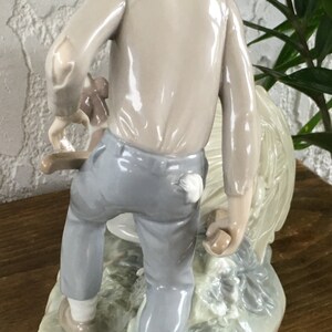 Puede incluir: Figura de cer&aacute;mica que representa a una persona con sombrero y ropa clara, que lleva un animal peque&ntilde;o. La figura es principalmente en tonos beige, gris y blanco, con una base detallada. La figura mide aproximadamente 15 cm de altura.