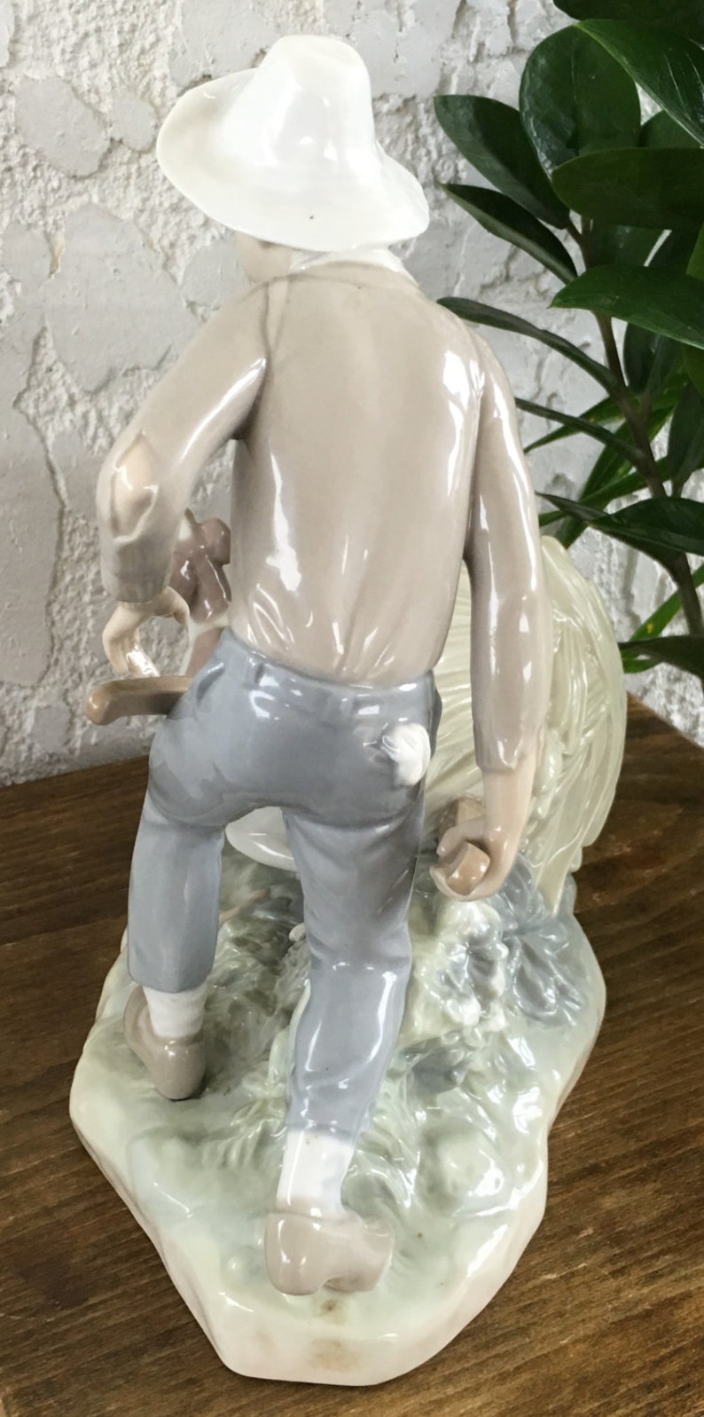 Puede incluir: Figura de cer&aacute;mica que representa a una persona con sombrero y ropa clara, que lleva un animal peque&ntilde;o. La figura es principalmente en tonos beige, gris y blanco, con una base detallada. La figura mide aproximadamente 15 cm de altura.