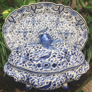 May include: A blue and white ceramic serving dish and lid with a floral and bird design. The dish has a scalloped edge and is decorated with intricate patterns. The lid features a bird-shaped handle.