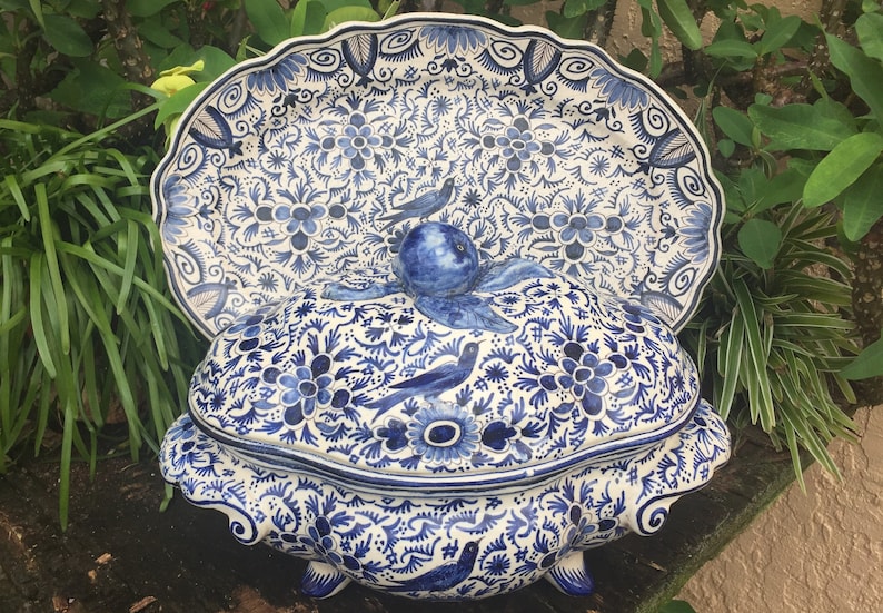 May include: A blue and white ceramic serving dish and lid with a floral and bird design. The dish has a scalloped edge and is decorated with intricate patterns. The lid features a bird-shaped handle.