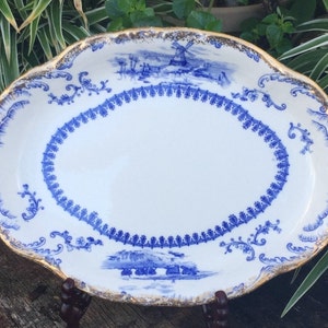May include: An oval ceramic platter featuring a white base with blue transferware patterns. The design includes a central oval border, a windmill landscape, and ornate scrollwork. The scalloped edges are trimmed with a worn gold metallic finish.