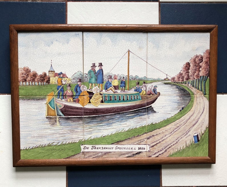 Puede incluir: Un azulejo vintage que representa una escena de canal con un barco lleno de pasajeros. El barco est&aacute; etiquetado como "De Trekschuit Omstreeks 1850". El azulejo est&aacute; enmarcado en madera.
