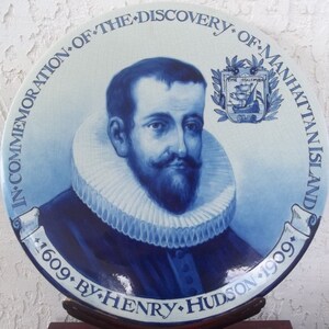K&ouml;nnte beinhalten: Ein blauer und wei&szlig;er Keramikteller, der an die Entdeckung von Manhattan Island durch Henry Hudson im Jahr 1609 erinnert. Der Teller zeigt ein Portr&auml;t von Hudson und ein Wappen mit einem Schiff.