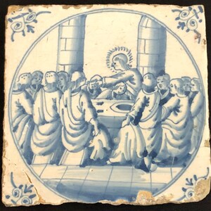 May include: A square, antique ceramic tile featuring a blue and white illustration of a religious scene. The image shows a group of figures gathered around a table, with one figure in the center. The tile has a circular border and decorative corner accents.