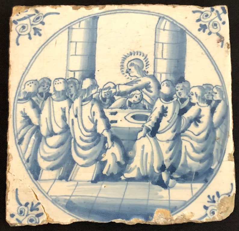 May include: A square, antique ceramic tile featuring a blue and white illustration of a religious scene. The image shows a group of figures gathered around a table, with one figure in the center. The tile has a circular border and decorative corner accents.
