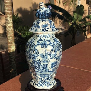 May include: A blue and white ceramic jar with a lid, featuring floral and figure designs. The jar has a small sculpture on top of the lid. The jar is displayed on a dark wooden stand.