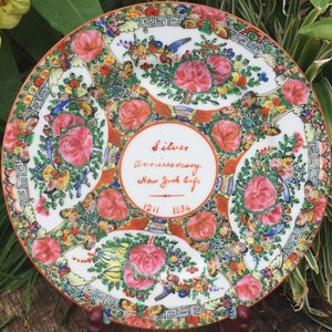 Op de afbeelding: Een wit porseleinen bord met een kleurrijk bloemenontwerp. Het bord heeft een rode rand en een centrale inscriptie met de tekst "Silver Anniversary New York Cafe 1911 1936".
