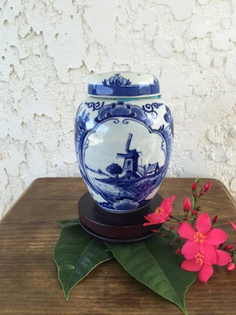 May include: A blue and white ceramic jar with a lid, featuring a windmill scene. The jar sits on a dark wooden base, with pink flowers and green leaves in the foreground. The jar is approximately 6 inches tall.
