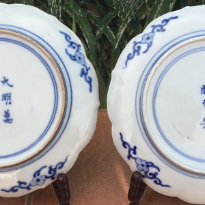 May include: Two antique white porcelain plates with scalloped edges and blue floral designs. Each plate has a blue ring around the center and blue text. The plates are displayed on a brown surface.