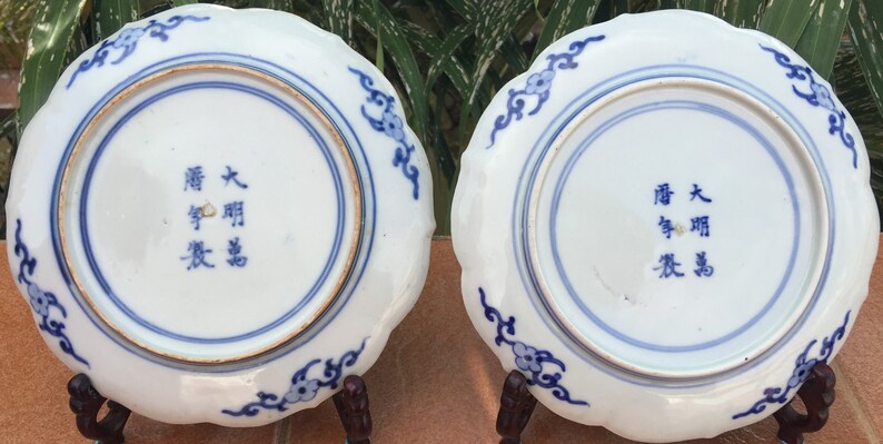 May include: Two antique white porcelain plates with scalloped edges and blue floral designs. Each plate has a blue ring around the center and blue text. The plates are displayed on a brown surface.