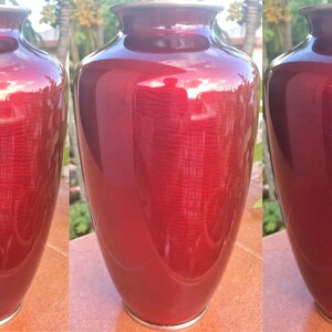 Puede incluir: Un jarr&oacute;n de cloisonn&eacute; rojo con un dise&ntilde;o floral blanco y dorado. El jarr&oacute;n est&aacute; decorado con una rosa estilizada y hojas.