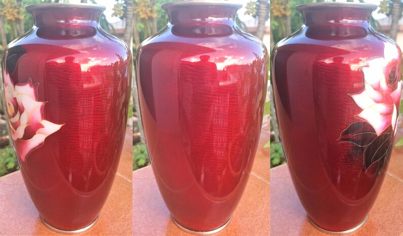 Puede incluir: Un jarr&oacute;n de cloisonn&eacute; rojo con un dise&ntilde;o floral blanco y dorado. El jarr&oacute;n est&aacute; decorado con una rosa estilizada y hojas.