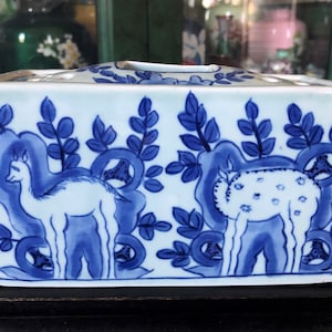 Op de afbeelding: Rechthoekige keramische vaas met een blauw en wit ontwerp. De vaas heeft een herhalend patroon van gestileerde herten en bladeren. De vaas is een decoratief item, waarschijnlijk voor bloemstukken.